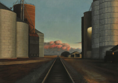 Railroad tracks run through the center of a landscape flanked by large grain silos at dusk, with glowing pink clouds on the horizon.