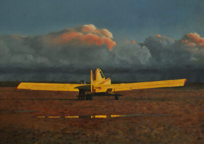 A bright yellow agricultural aircraft parked on a dark, wet airfield before a backdrop of heavy, storm-lit clouds.